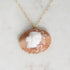 A gold-tone necklace featuring an oval carved shell pendant with a white classical bust profile on a warm peach-brown background, displayed on a light marble surface.