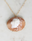 A gold-tone necklace featuring an oval carved shell pendant with a white classical bust profile on a warm peach-brown background, displayed on a light marble surface.