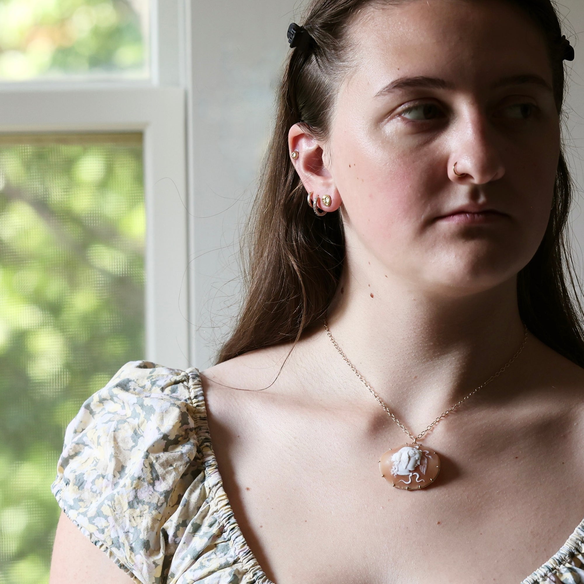 A close-up lifestyle shot of a young woman wearing a gold-toned necklace with a cameo-style pendant and matching stud earrings, set against a soft-focus background of a window.