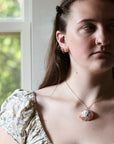 A close-up lifestyle shot of a young woman wearing a gold-toned necklace with a cameo-style pendant and matching stud earrings, set against a soft-focus background of a window.