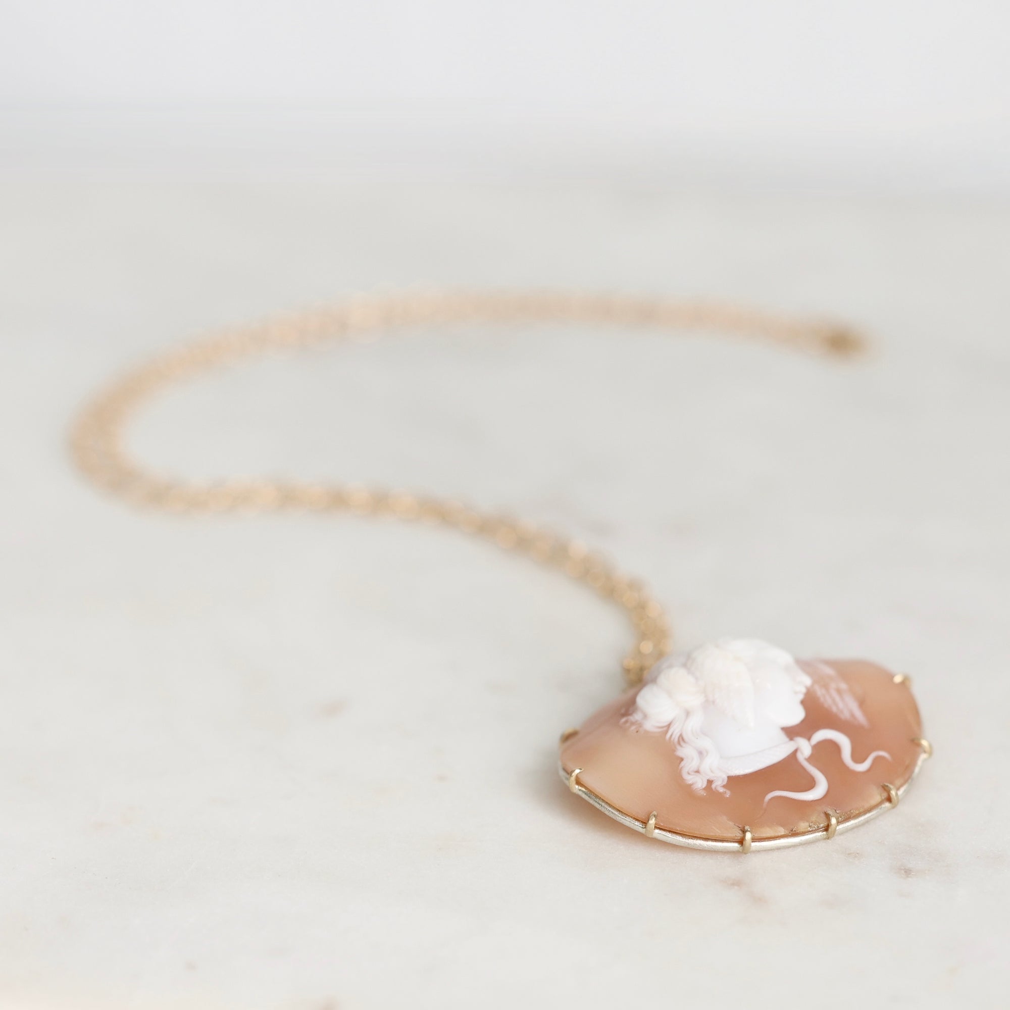 A gold necklace with a carved cameo pendant on a peach-colored shell, resting on a light marble surface.