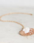 A gold necklace with a carved cameo pendant on a peach-colored shell, resting on a light marble surface.