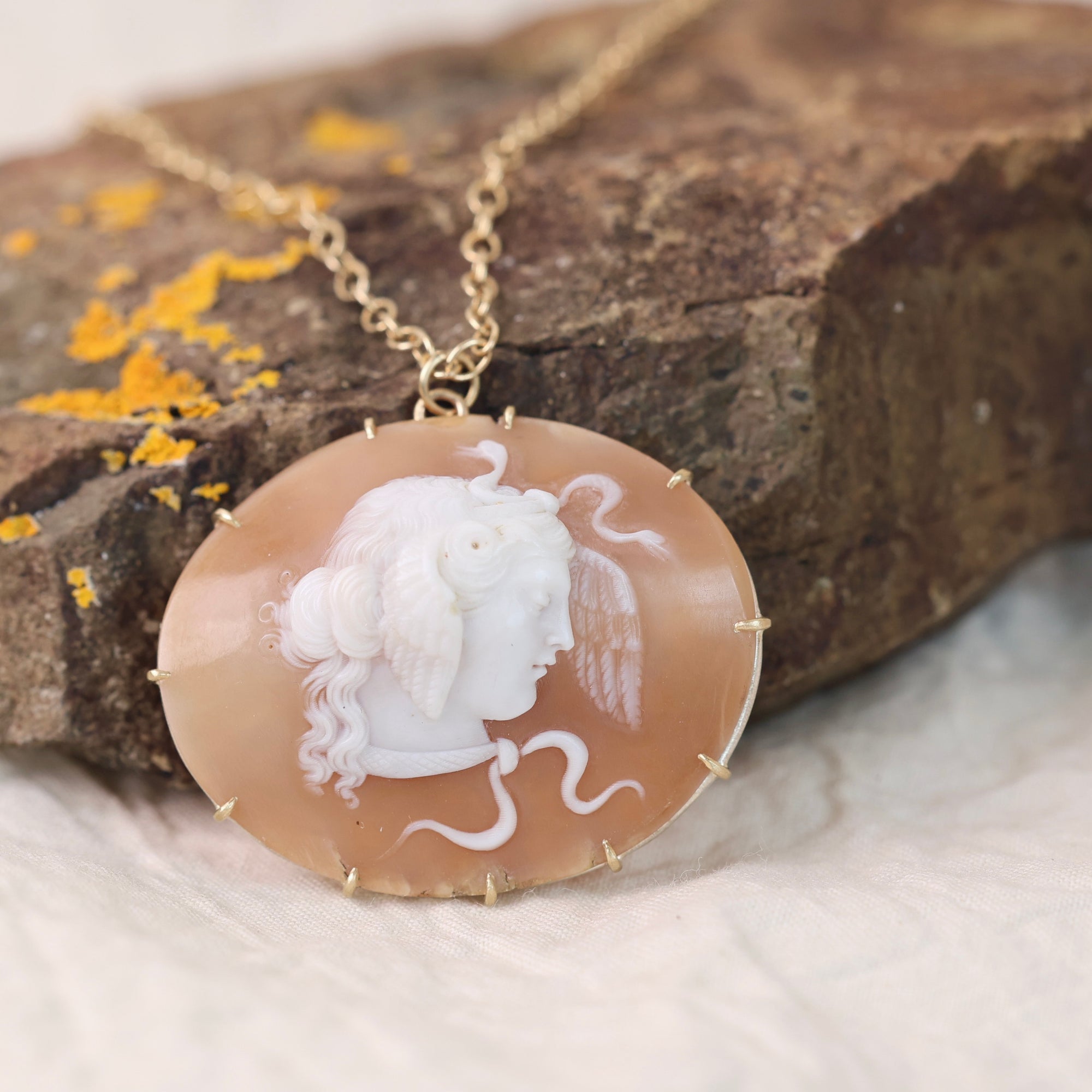 A gold-tone necklace with an oval carved pendant featuring a white classical profile on a peach-orange translucent stone, displayed on a textured brown rock with small yellow lichen accents.