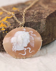 A gold-tone necklace with an oval carved pendant featuring a white classical profile on a peach-orange translucent stone, displayed on a textured brown rock with small yellow lichen accents.
