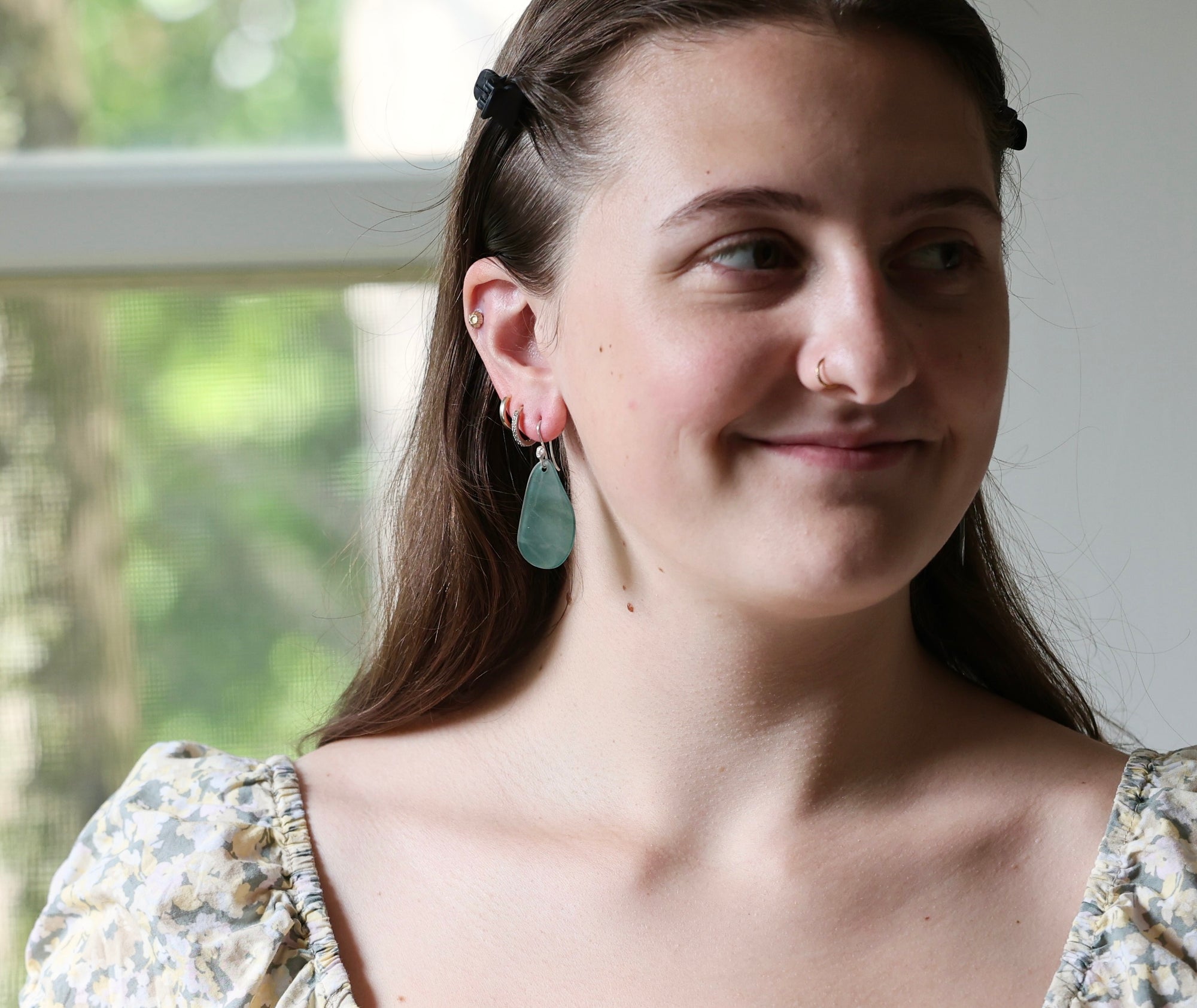 A close-up lifestyle shot of a young woman with brown hair wearing a small gold hoop earring and a light green teardrop dangle earring. She is smiling and looking to the side, wearing a floral-patterned top against a softly blurred indoor background.