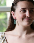 A close-up lifestyle shot of a young woman with brown hair wearing a small gold hoop earring and a light green teardrop dangle earring. She is smiling and looking to the side, wearing a floral-patterned top against a softly blurred indoor background.