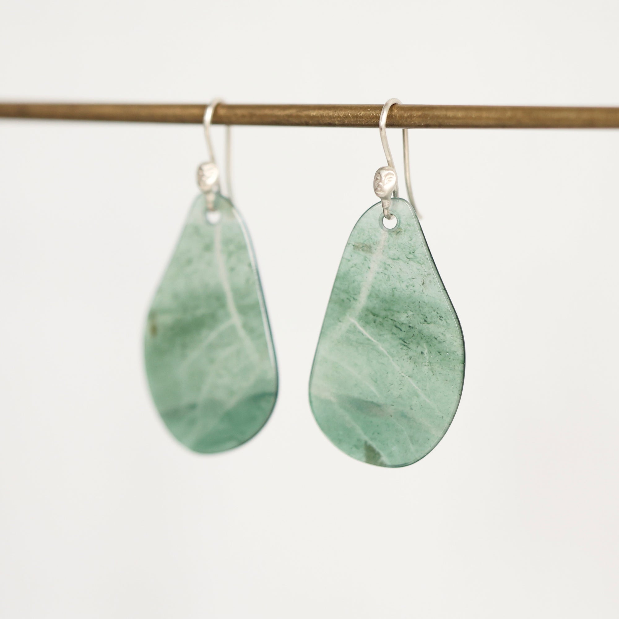 A pair of teardrop-shaped green stone pendants with silver fishhook ear wires, hanging from a thin horizontal wooden rod against a plain light background.