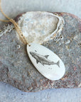 A gold chain necklace with an oval white pendant featuring a detailed black engraving of a shark, displayed on a textured gray rock.