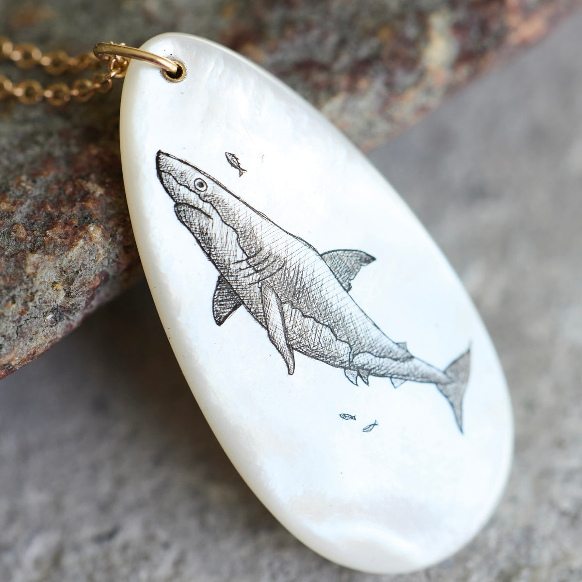A white mother-of-pearl pendant necklace featuring a detailed black ink sketch of a shark, hanging from a gold chain and resting on a textured rock.