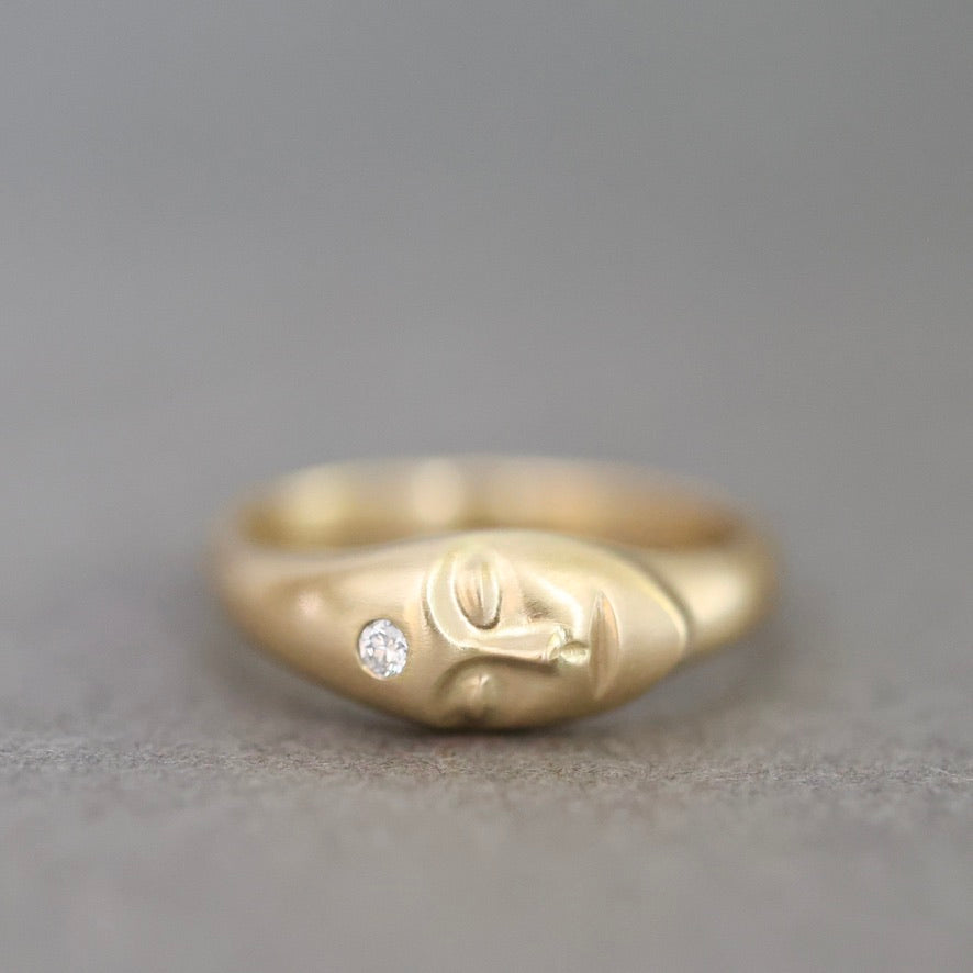 A gold ring featuring a stylized head design with a small clear gemstone on the side, set against a neutral gray background.