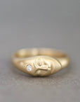 A gold ring featuring a stylized head design with a small clear gemstone on the side, set against a neutral gray background.