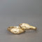 A pair of gold rings shaped like stylized faces, resting on a neutral gray surface. One ring features a small clear gemstone.