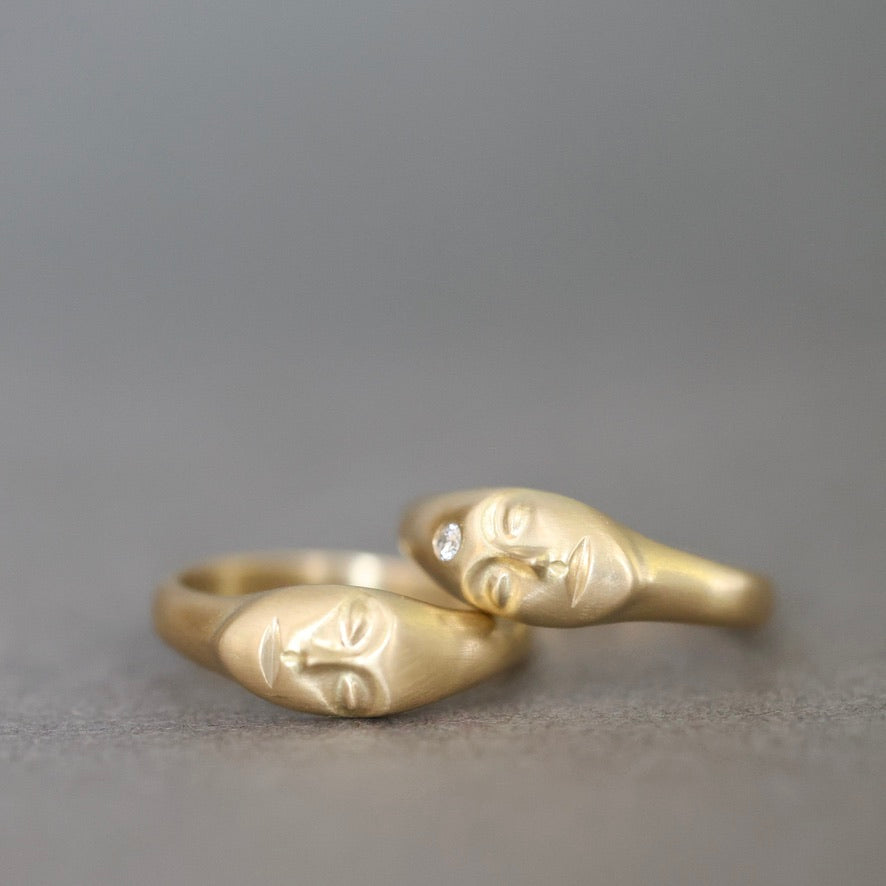 A pair of gold rings shaped like stylized faces, resting on a neutral gray surface. One ring features a small clear gemstone.
