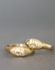 A pair of gold rings shaped like stylized faces, resting on a neutral gray surface. One ring features a small clear gemstone.