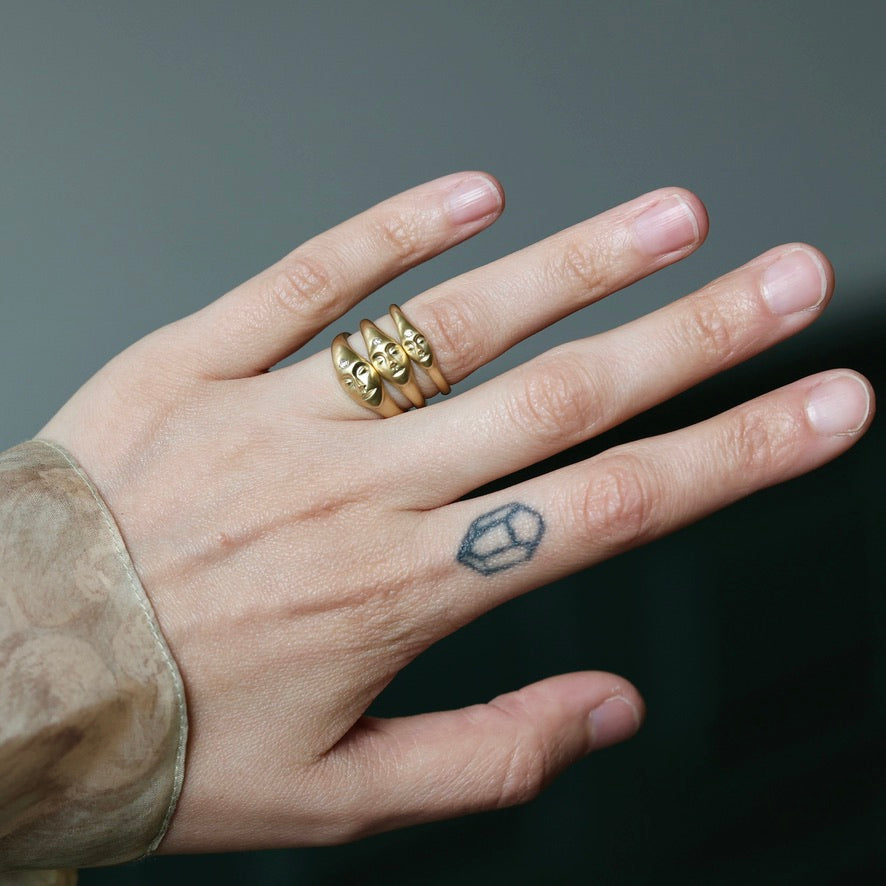 A close-up of a hand wearing three gold rings, with a small black geometric tattoo on the ring finger. The background is a dark, neutral gray.