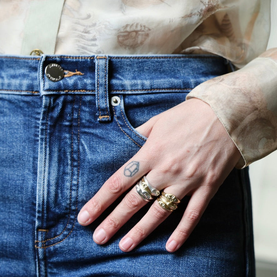 A close-up of a person's hand resting on the pocket of blue denim jeans, wearing three rings: a silver ring with a small rectangular stone, a textured silver band, and a gold ring with a detailed sculpted design.