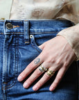 A close-up of a person's hand resting on the pocket of blue denim jeans, wearing three rings: a silver ring with a small rectangular stone, a textured silver band, and a gold ring with a detailed sculpted design.
