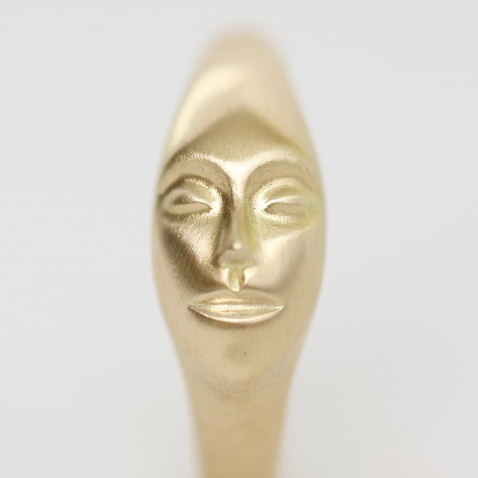 A close-up of a metallic gold object with a stylized human face sculpted on it, shown against a plain light gray background.