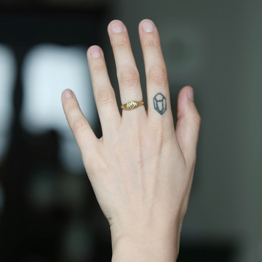 A close-up of a person's hand showing two rings: a small gold band ring on the ring finger and a tiny dark geometric tattoo or sticker on the middle finger, against a blurred dark background.