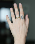 A close-up of a person's hand showing two rings: a small gold band ring on the ring finger and a tiny dark geometric tattoo or sticker on the middle finger, against a blurred dark background.