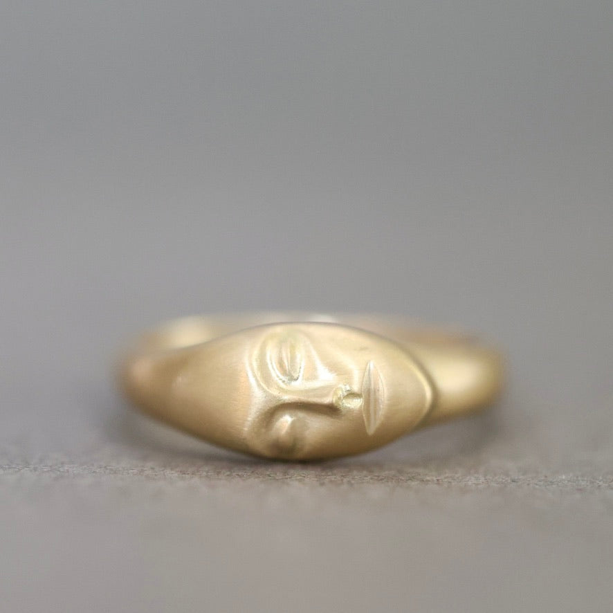A gold ring sculpted in the shape of a stylized face, photographed against a neutral gray background.
