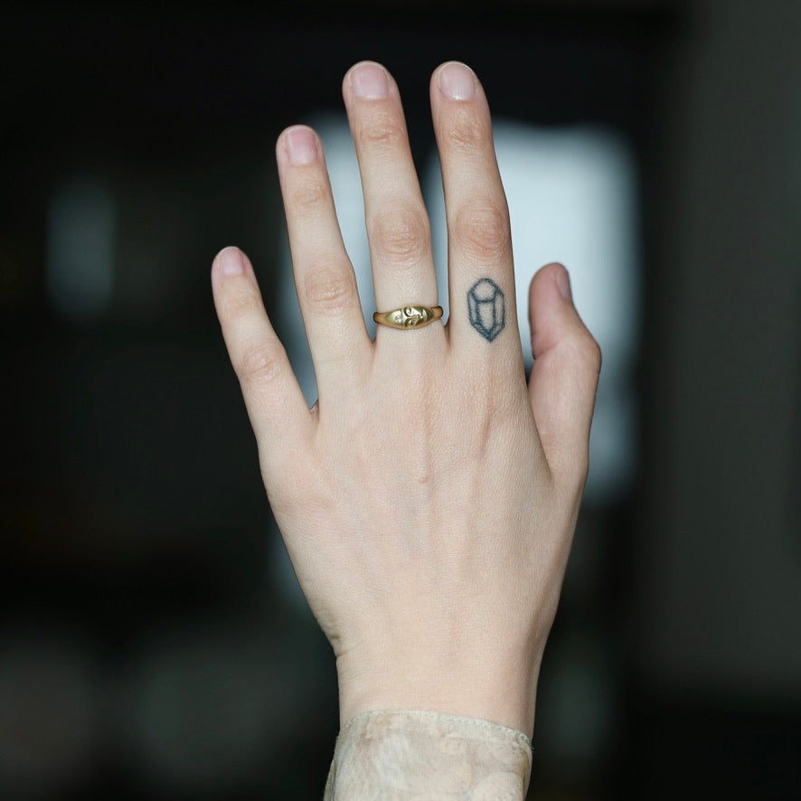 A close-up of a person's hand wearing a gold signet ring with a small geometric emblem, with a small geometric tattoo on the middle finger, against a dark blurred background.