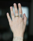 A close-up of a person's hand wearing a gold signet ring with a small geometric emblem, with a small geometric tattoo on the middle finger, against a dark blurred background.