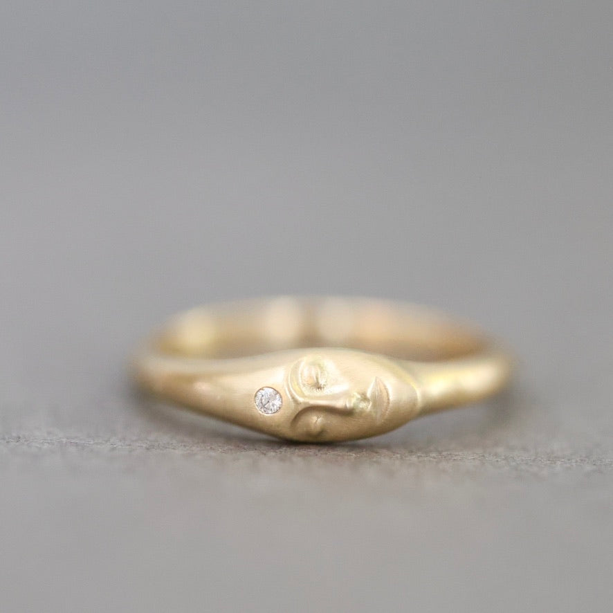 A gold ring featuring a sculpted face with a small clear gemstone on its forehead, photographed on a neutral gray background.