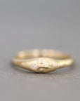 A gold ring featuring a sculpted face with a small clear gemstone on its forehead, photographed on a neutral gray background.