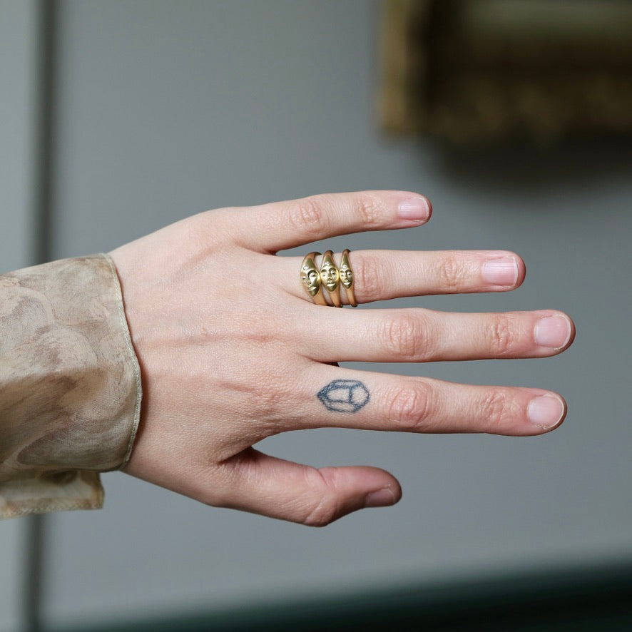 A close-up of a person's hand wearing three stacked gold rings, with a small geometric tattoo on the ring finger, set against a blurred gray background.