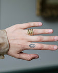 A close-up of a person's hand wearing three stacked gold rings, with a small geometric tattoo on the ring finger, set against a blurred gray background.