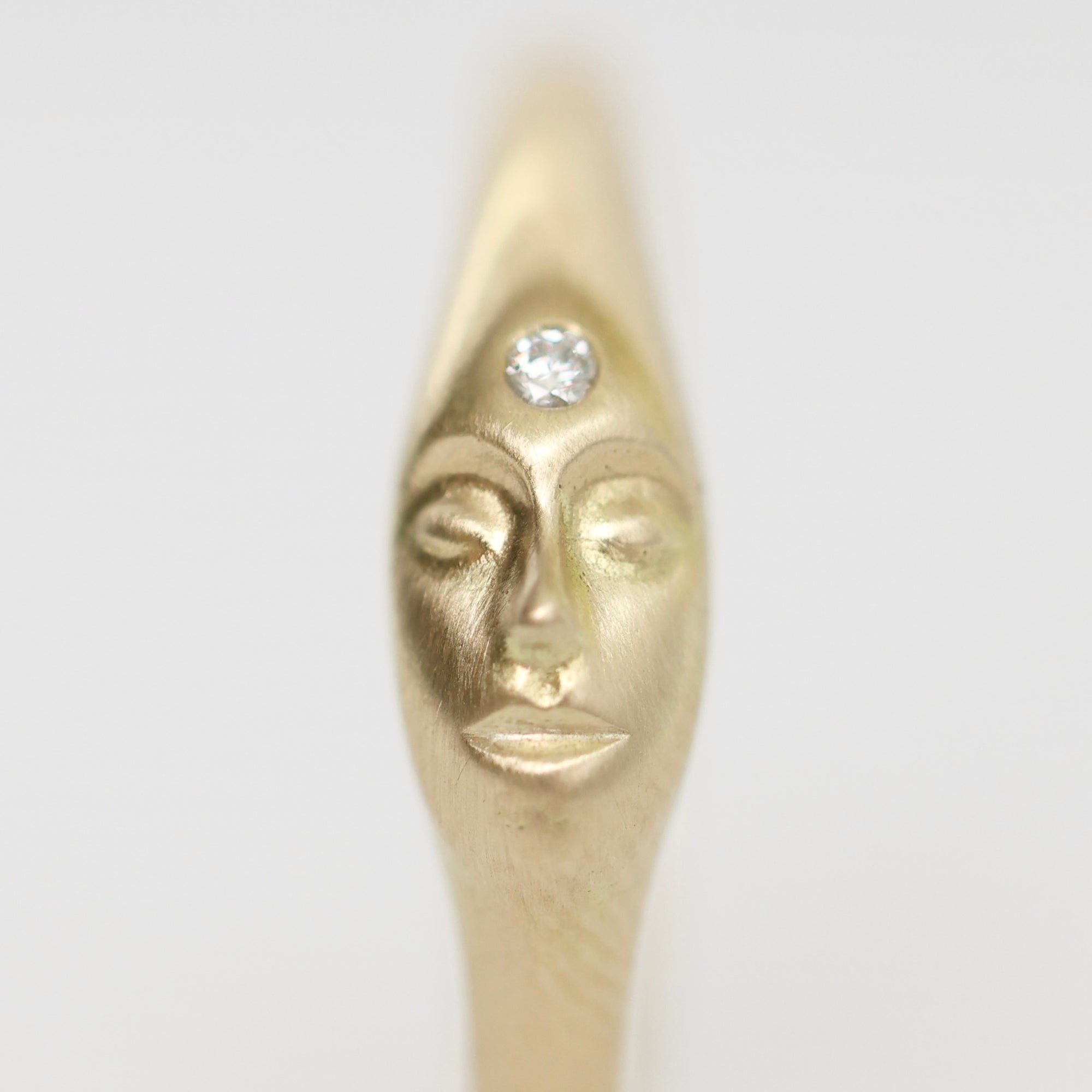 A gold-toned ring featuring a sculpted stylized face with closed eyes and a clear round gemstone set on the forehead, shown against a plain light gray background.