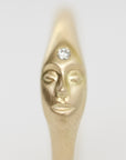 A gold-toned ring featuring a sculpted stylized face with closed eyes and a clear round gemstone set on the forehead, shown against a plain light gray background.