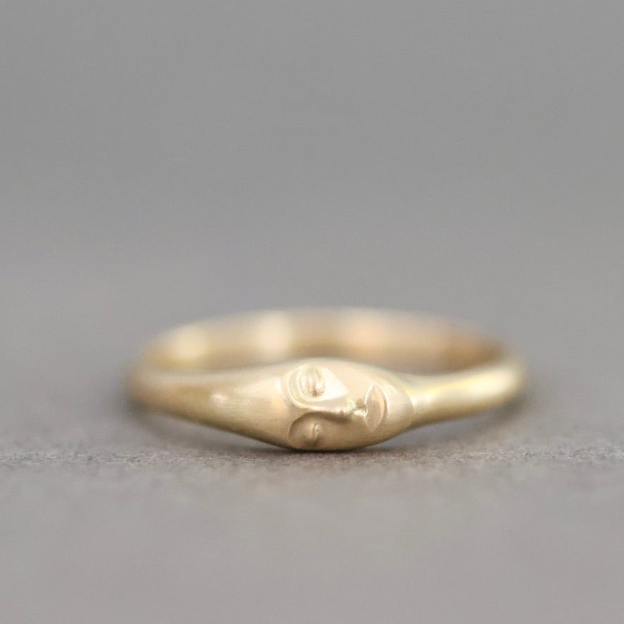 A small gold ring with a sculpted face motif on the band, photographed against a neutral gray background.