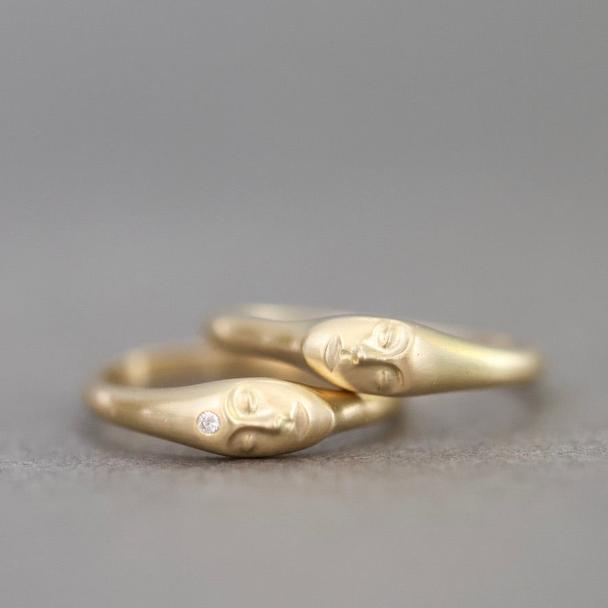 Two gold rings with sculpted faces resting on each other, one featuring a small clear gemstone, photographed on a neutral gray background.