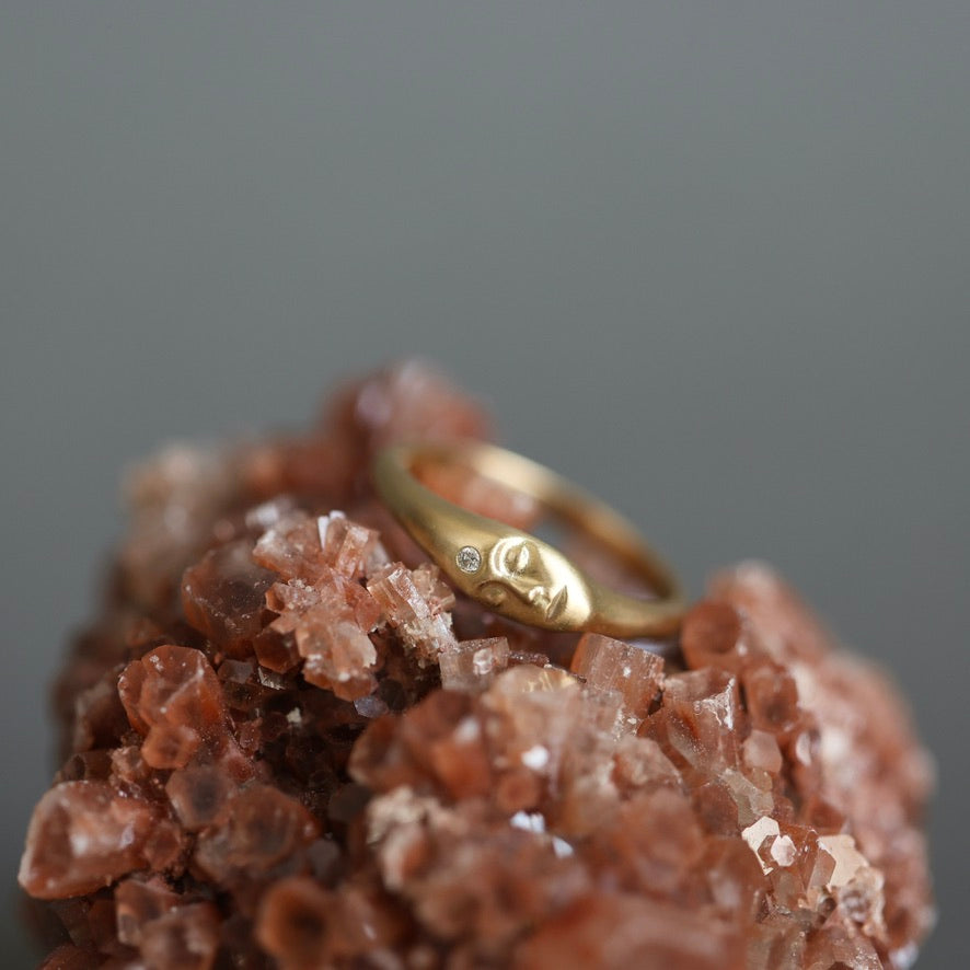 A gold ring with a small engraved face motif and a tiny clear stone, displayed on a cluster of brown and pink crystals against a soft gray background.
