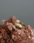 A gold ring with a small engraved face motif and a tiny clear stone, displayed on a cluster of brown and pink crystals against a soft gray background.