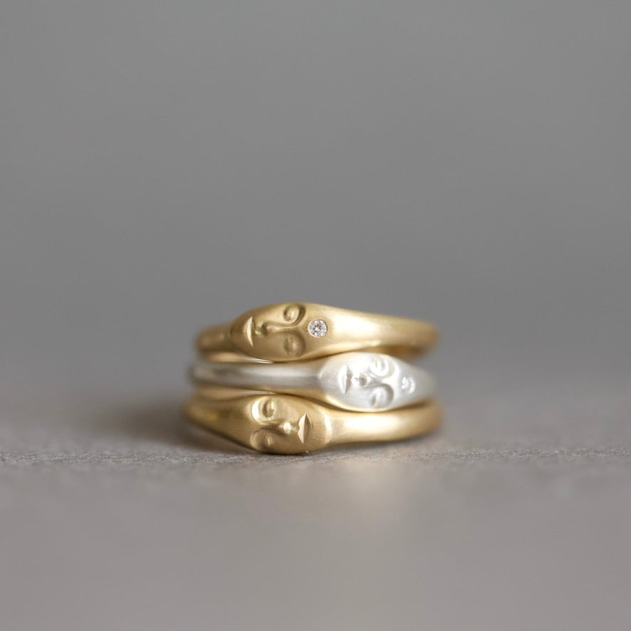 Three stacked rings featuring stylized human face profiles, with two in polished yellow gold and one in silver, set against a soft gray background.
