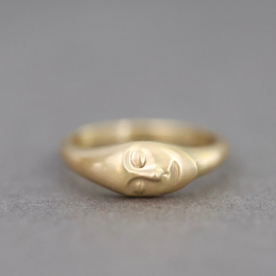 A gold ring featuring a stylized, abstract face design on the front, photographed against a neutral gray background.