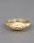 A gold ring featuring a stylized, abstract face design on the front, photographed against a neutral gray background.