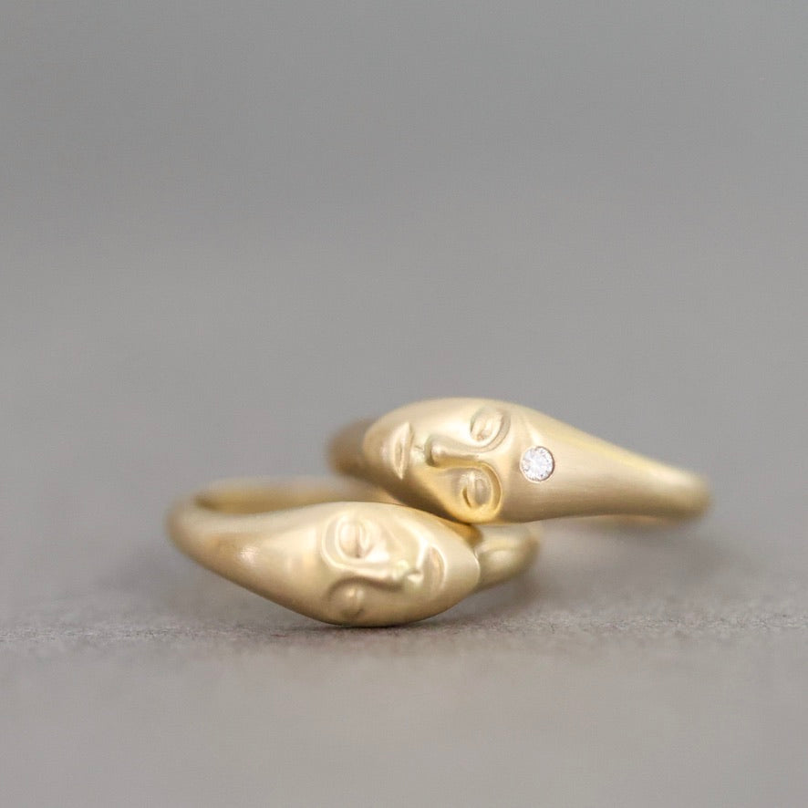 A gold ring featuring two sculpted faces facing each other, with a small clear gemstone set on the right face, photographed against a neutral gray background.