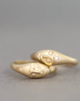 A gold ring featuring two sculpted faces facing each other, with a small clear gemstone set on the right face, photographed against a neutral gray background.
