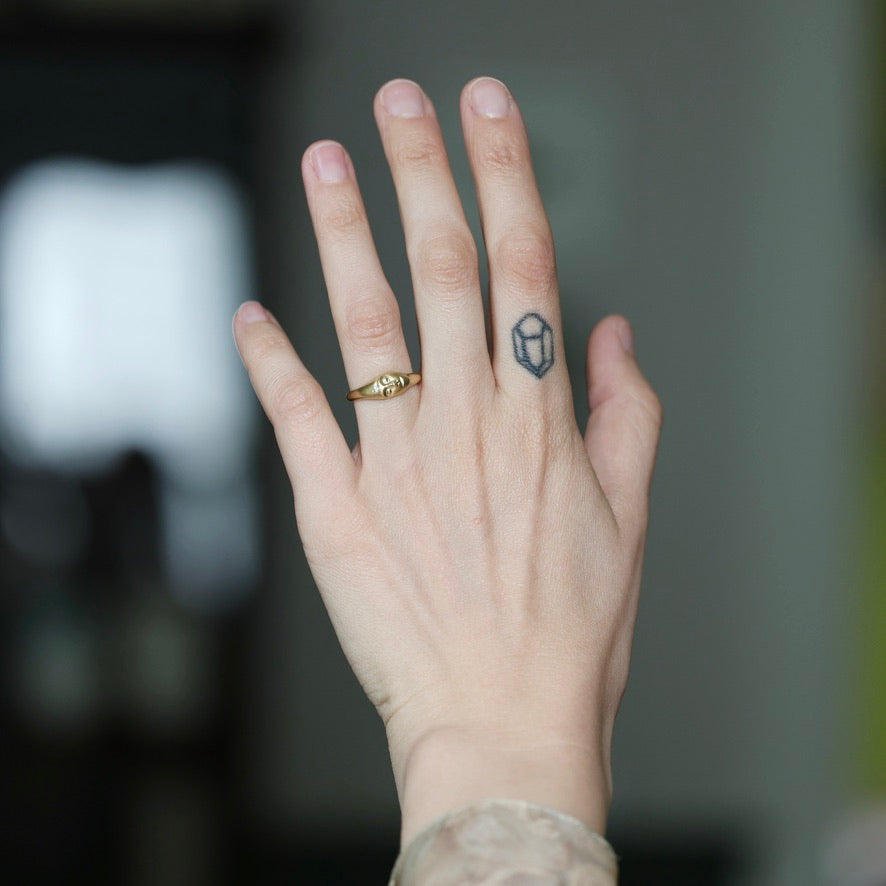 A close-up of a person's hand showing a small geometric diamond-shaped ring on the ring finger, with a tiny dark tattoo on the side of the finger, set against a blurred dark background.