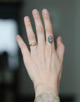 A close-up of a person's hand showing a small geometric diamond-shaped ring on the ring finger, with a tiny dark tattoo on the side of the finger, set against a blurred dark background.