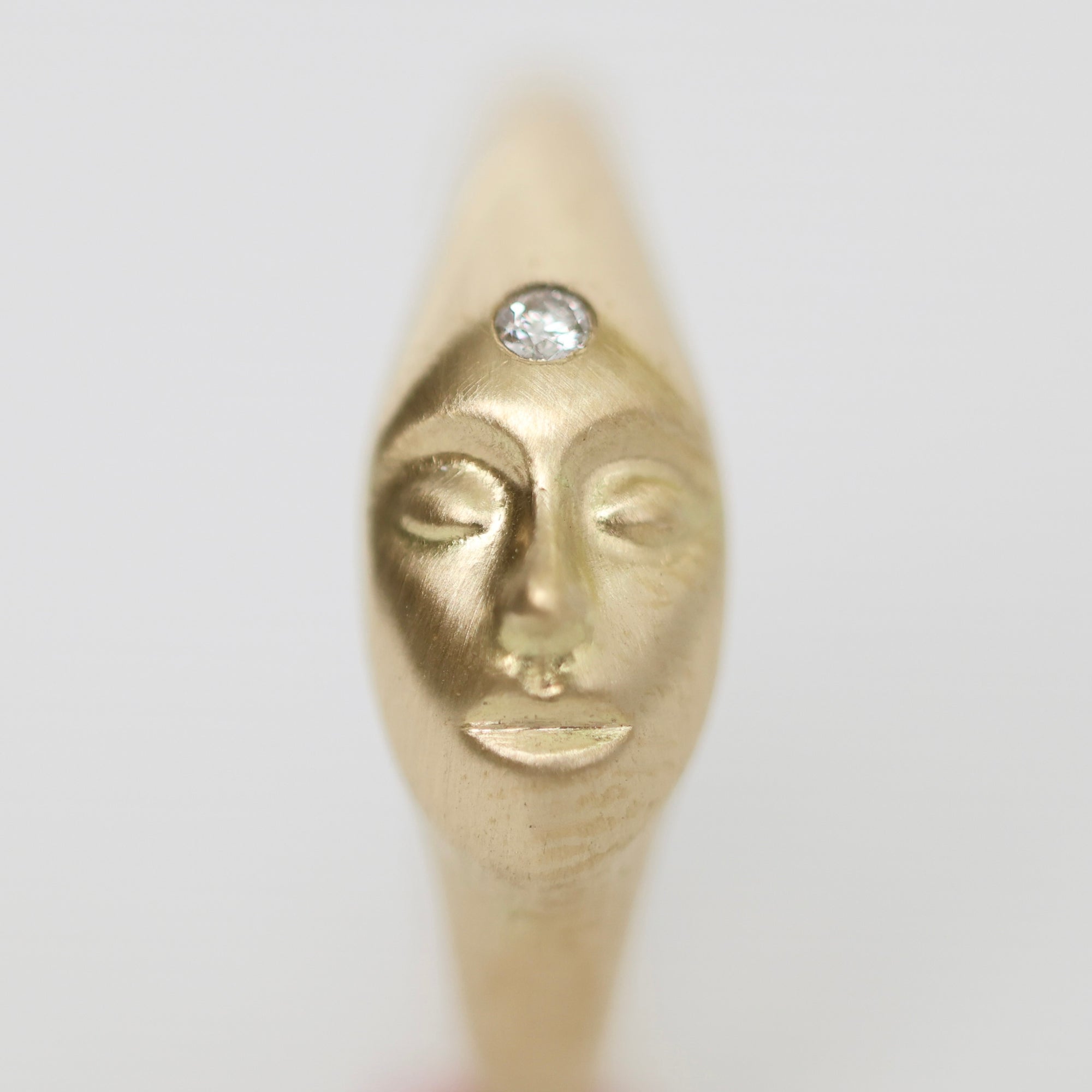 A gold-toned ring featuring a sculpted face with closed eyes and a clear round gemstone set on the forehead, shown against a plain light gray background.