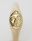 A gold-toned ring featuring a sculpted face with closed eyes and a clear round gemstone set on the forehead, shown against a plain light gray background.