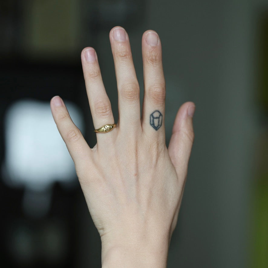 A close-up of a person's hand with a small geometric tattoo on the ring finger and a gold ring on the middle finger, set against a blurred dark background.