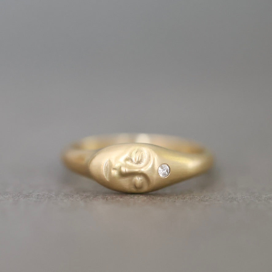 A gold ring featuring a stylized face design with a small clear gemstone on the side, photographed against a neutral gray background.