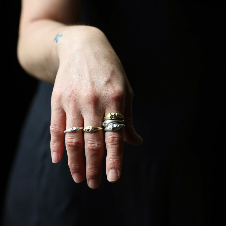 A close-up of a person's hand wearing several rings, including a silver band with a small gold oval accent and a silver band with a small gold oval accent, against a dark background.