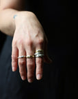 A close-up of a person's hand wearing several rings, including a silver band with a small gold oval accent and a silver band with a small gold oval accent, against a dark background.